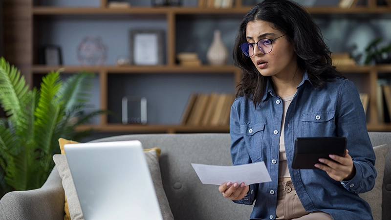 Worried woman with documents and a calculator looking at her laptop in disbelief.