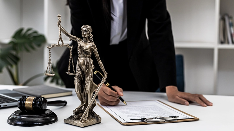 Attorney signing legal paperwork beside a Lady Justice statue and gavel.