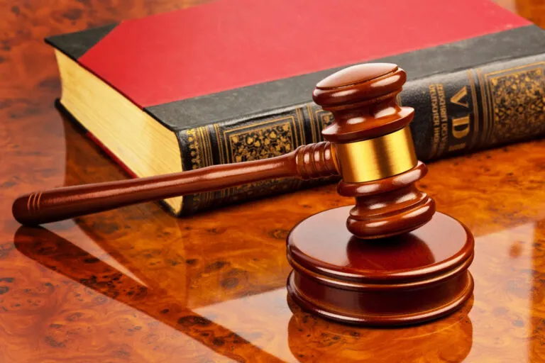 Wooden judge's gavel resting on a sound block next to a large red law book on a polished wooden desk.