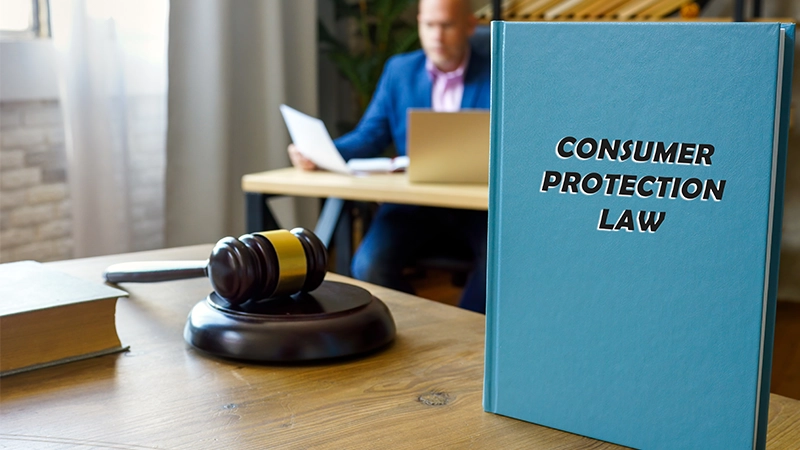 Blue "Consumer Protection Law" book and gavel on a desk, with a blurred lawyer in the background.