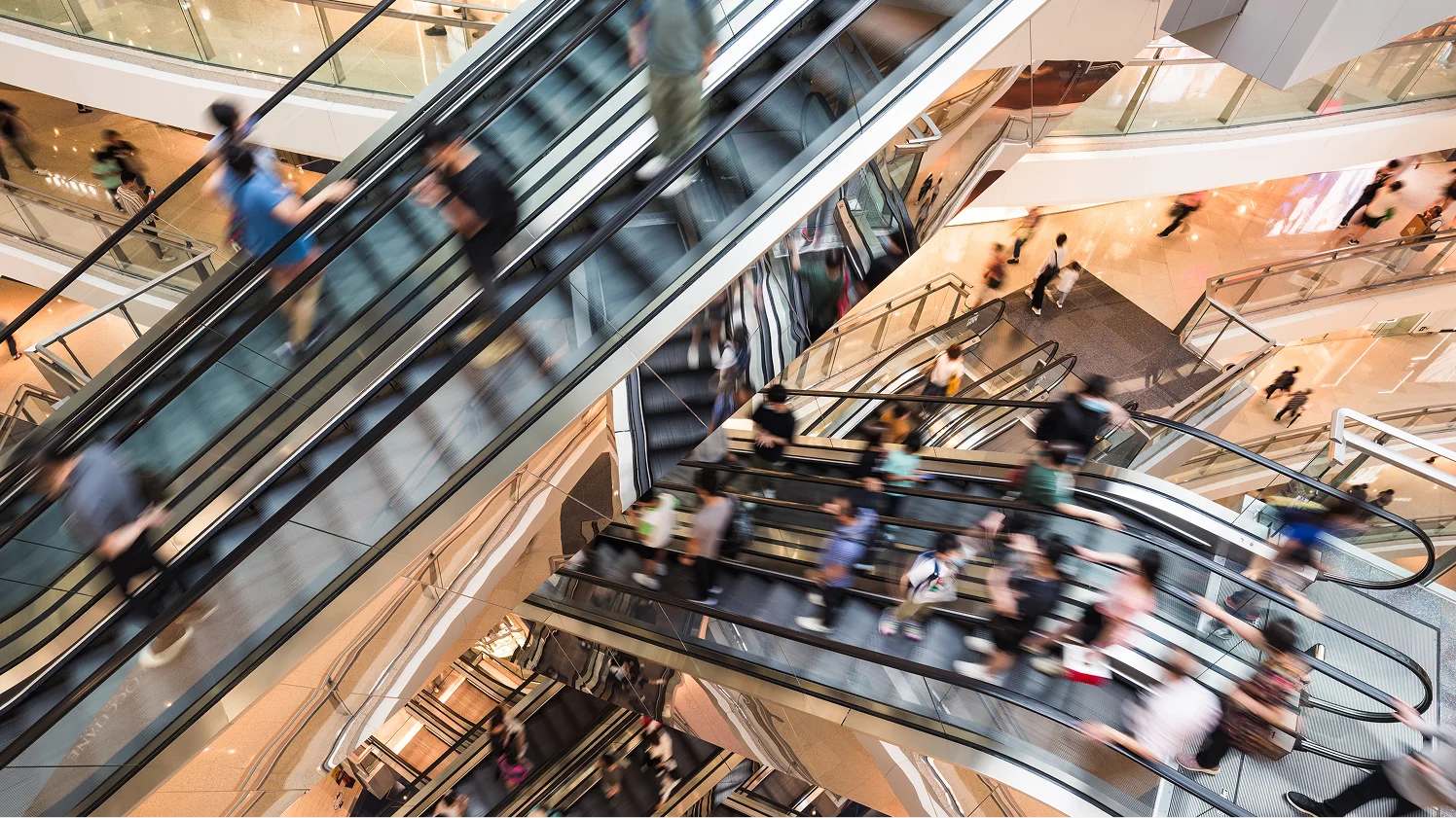 Blurred view of people riding multiple escalators inside a multi-level shopping mall.