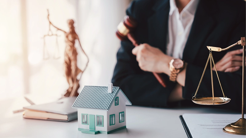 A female real estate lawyer in a suit holds a house model and scales of justice, signifying legal consulting.