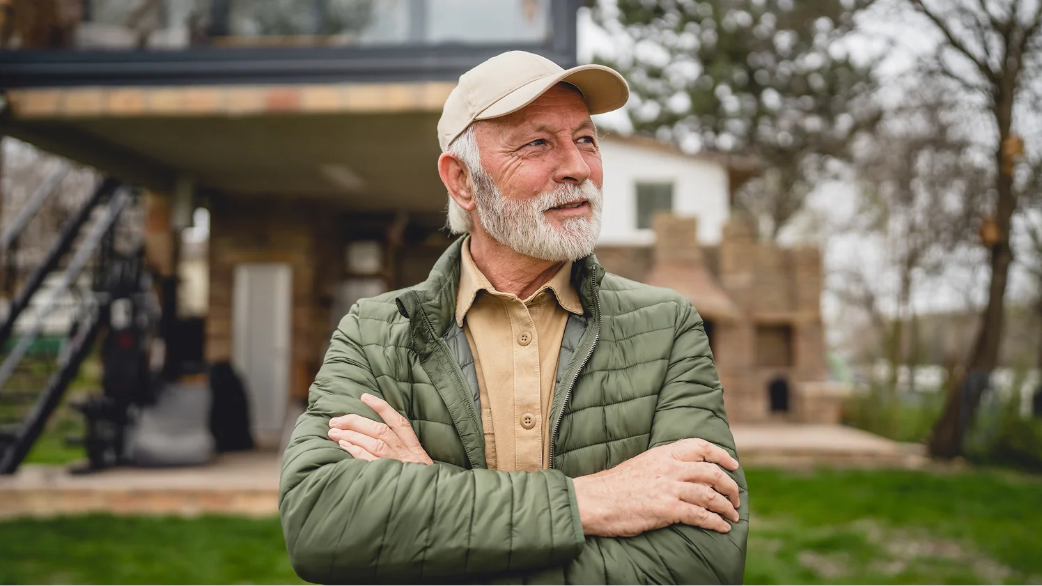 Senior man with a white beard wearing a baseball cap and green jacket standing outdoors with arms crossed.