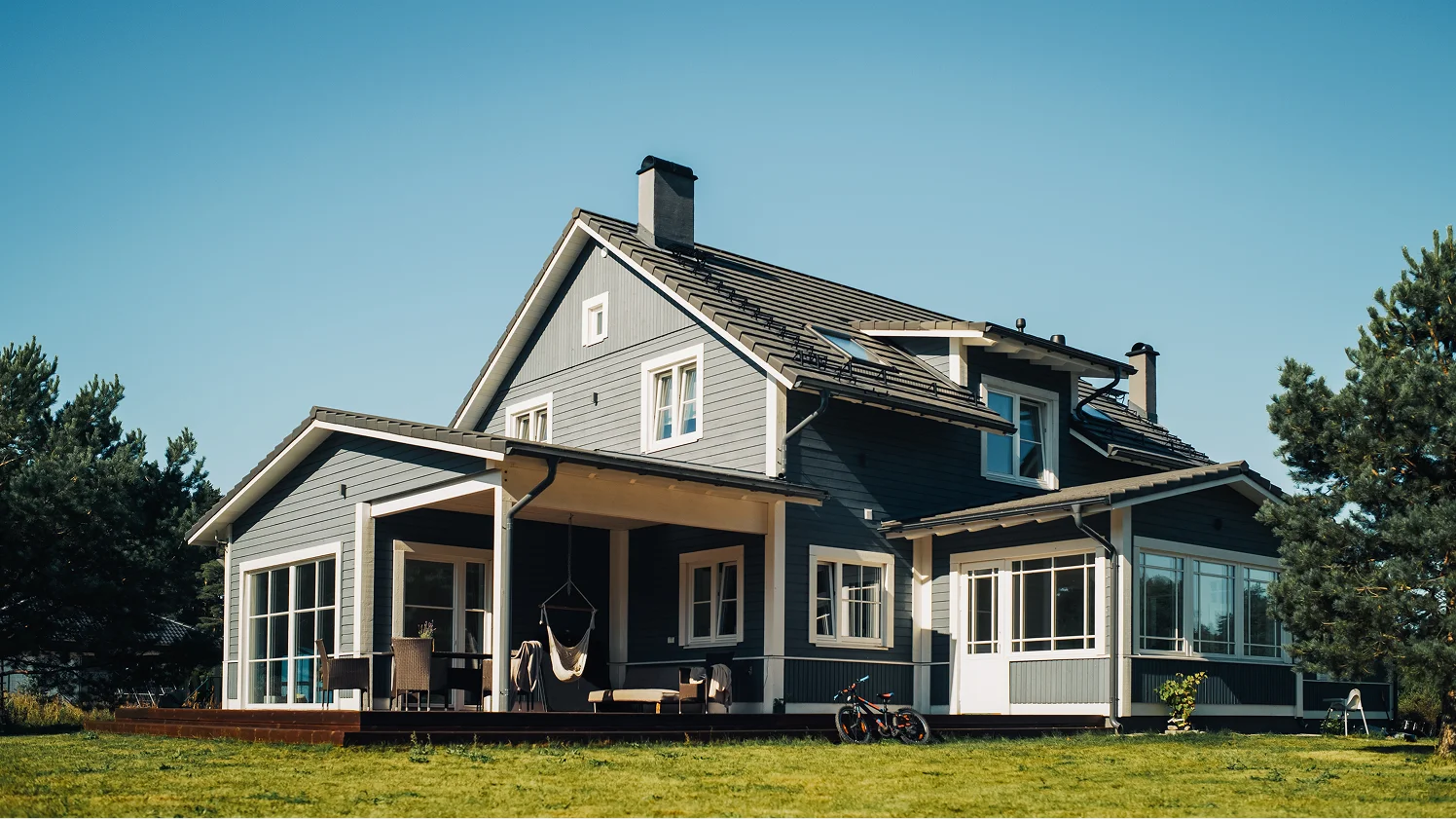 Two-story grey wooden house on a grassy lawn under a blue sky.