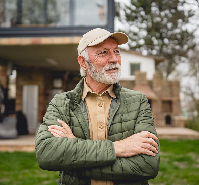 Senior man with a white beard wearing a baseball cap and green jacket standing outdoors with arms crossed.