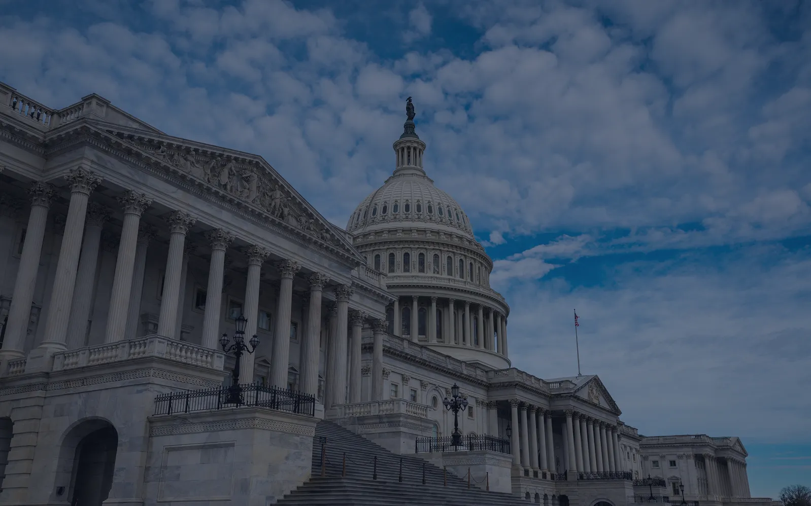 Low-angle view of the United States Capitol dome and classical facade against a partly cloudy blue sky.