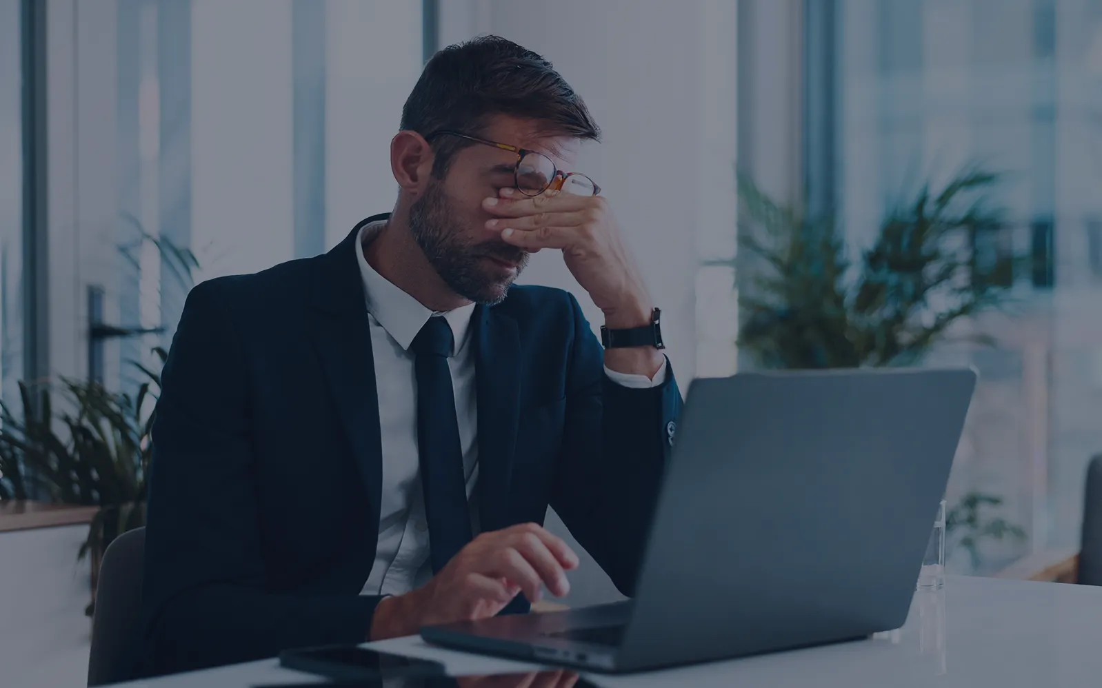 Stressed businessman in a suit sitting at his office desk with his hand covering his face while working on a laptop.